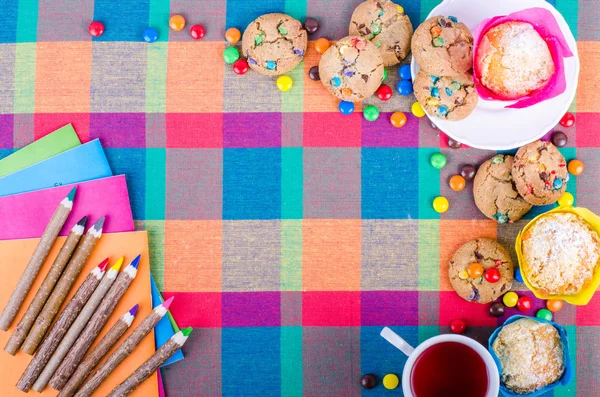 Свежие домашние кексы и печенье с цветной шоколадные конфеты с покрытием, красочные деревянные карандаши и тетради, тетради, чашка чая на клетчатой ​​кухонным полотенцем. Свободное пространство для вашего текста — стоковое фото